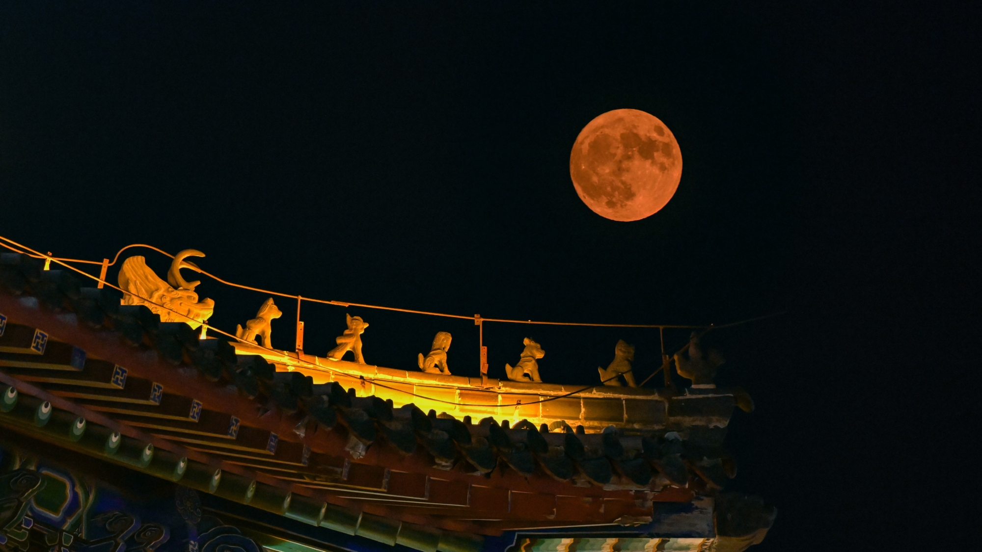 a full moon shines an orange rusty hue above an ornate rooftop decorated with colorful tiles and animal figures.
