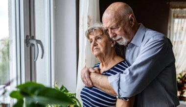 A photo of a senior man hugging his wife in front of a window in their home.
