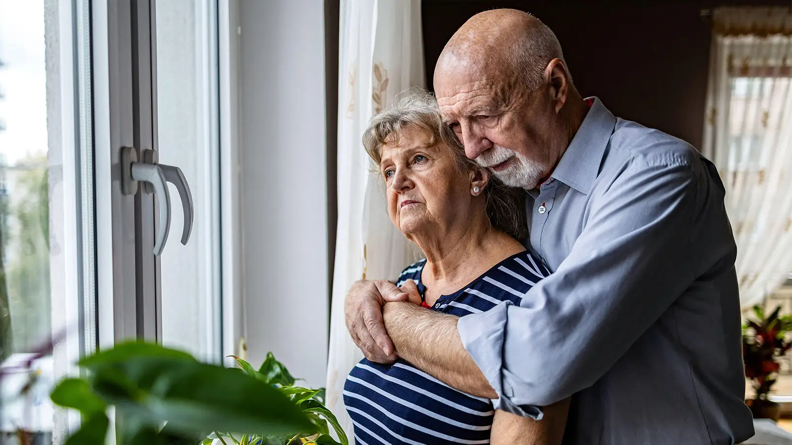 A photo of a senior man hugging his wife in front of a window in their home.