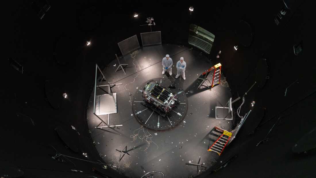 Downward-looking view of two people in white clean suits standing next to a spacecraft at the bottom of a large black circular chamber. The spacecraft lies at center on a black circular plate crisscrossed with white lines. Around the perimeter of the chamber are ladders and various stands. Several small lamps near the chamber’s floor create pools of light. There is a door open to the outside of the chamber at about 1 o’clock.
