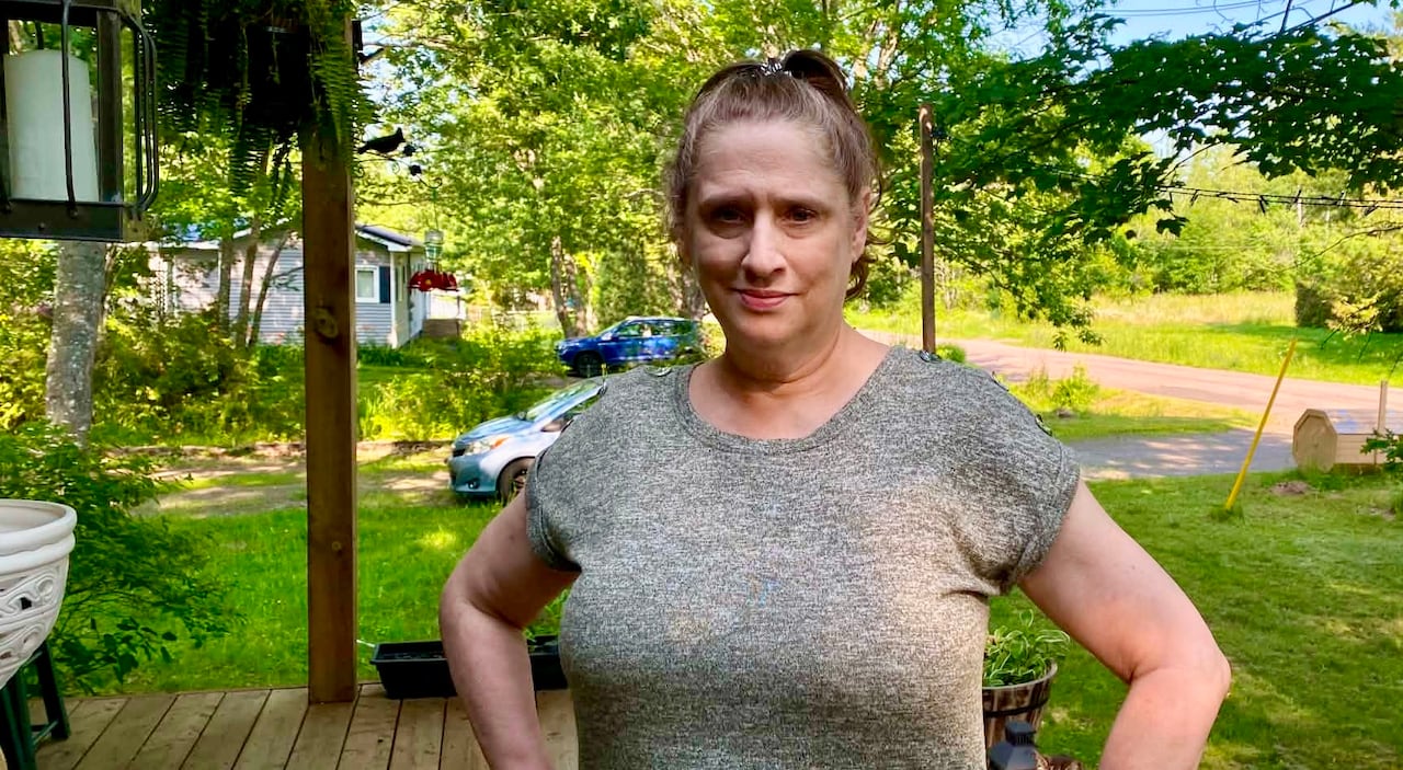 A woman with brown hair in a high ponytail looks at the camera while standing on a front porch in summer, with her hands on her hips.