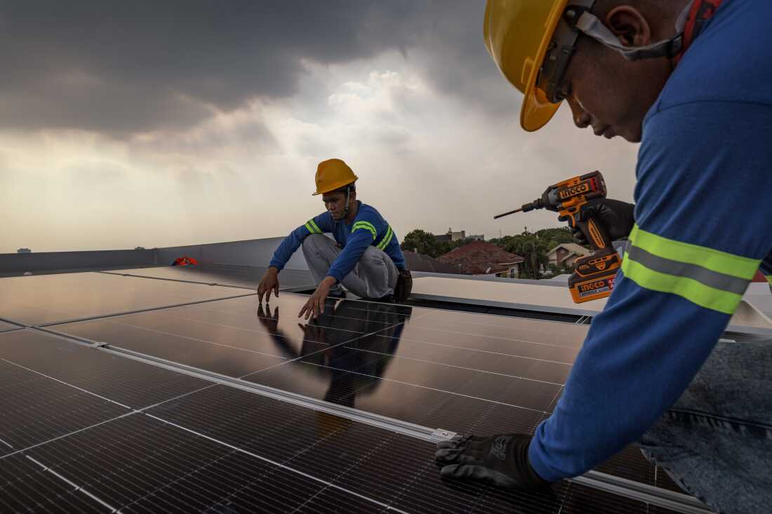 Workers wearing hard hats install solar panels on the roof of a house under a sky filled with gray clouds.