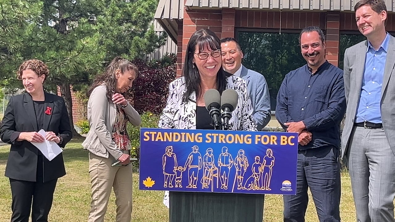 A woman with dark hair and glasses smiles as she stands in front of a podium on a sunny day in Prince George.