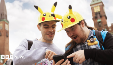 Two Pokémon Go fans wearing hats shaped like the yellow Pokémon character Pikachu are pointing and looking at a phone, in shock.