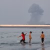Three boys play in the shallow waters of the Strait of Hormuz, as a plume of smoke rises from an explosion in the background, off Bandar Abbas, Iran, Monday.