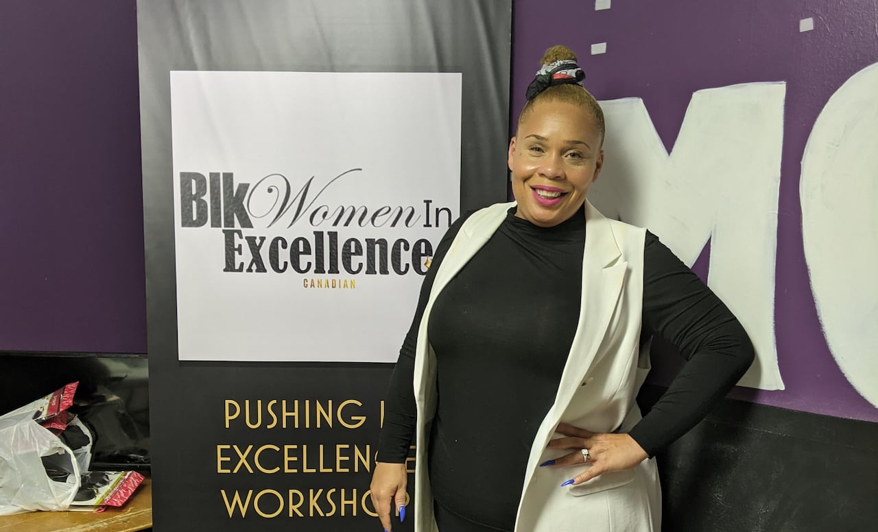A Black woman standing beside a sign that reads "Blk Women in Excellence"