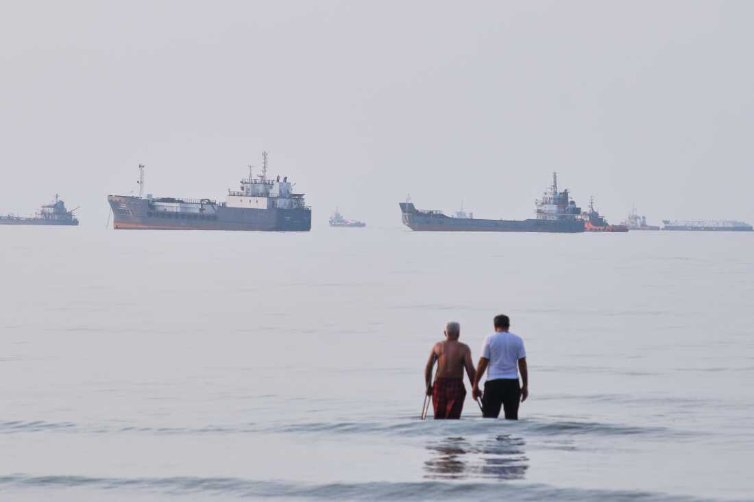 Two men wade in the waters of the Strait of Hormuz with vessels anchored in the background, off Bandar Abbas, Iran, Sunday, July 12, 2026.