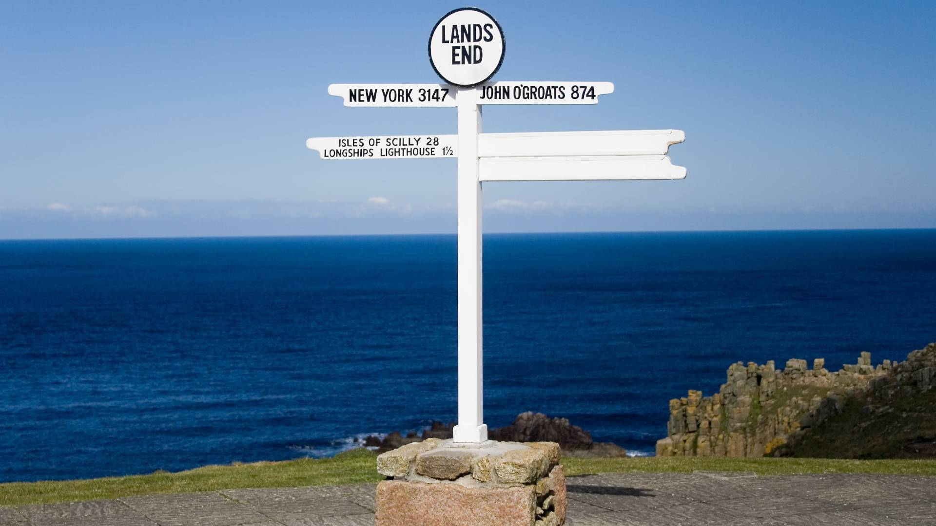 White signpost at Land's End, England, shows distances to New York, John O'Groats, Isles of Scilly, and Longships Lighthouse, with blue sea and sky in the background.