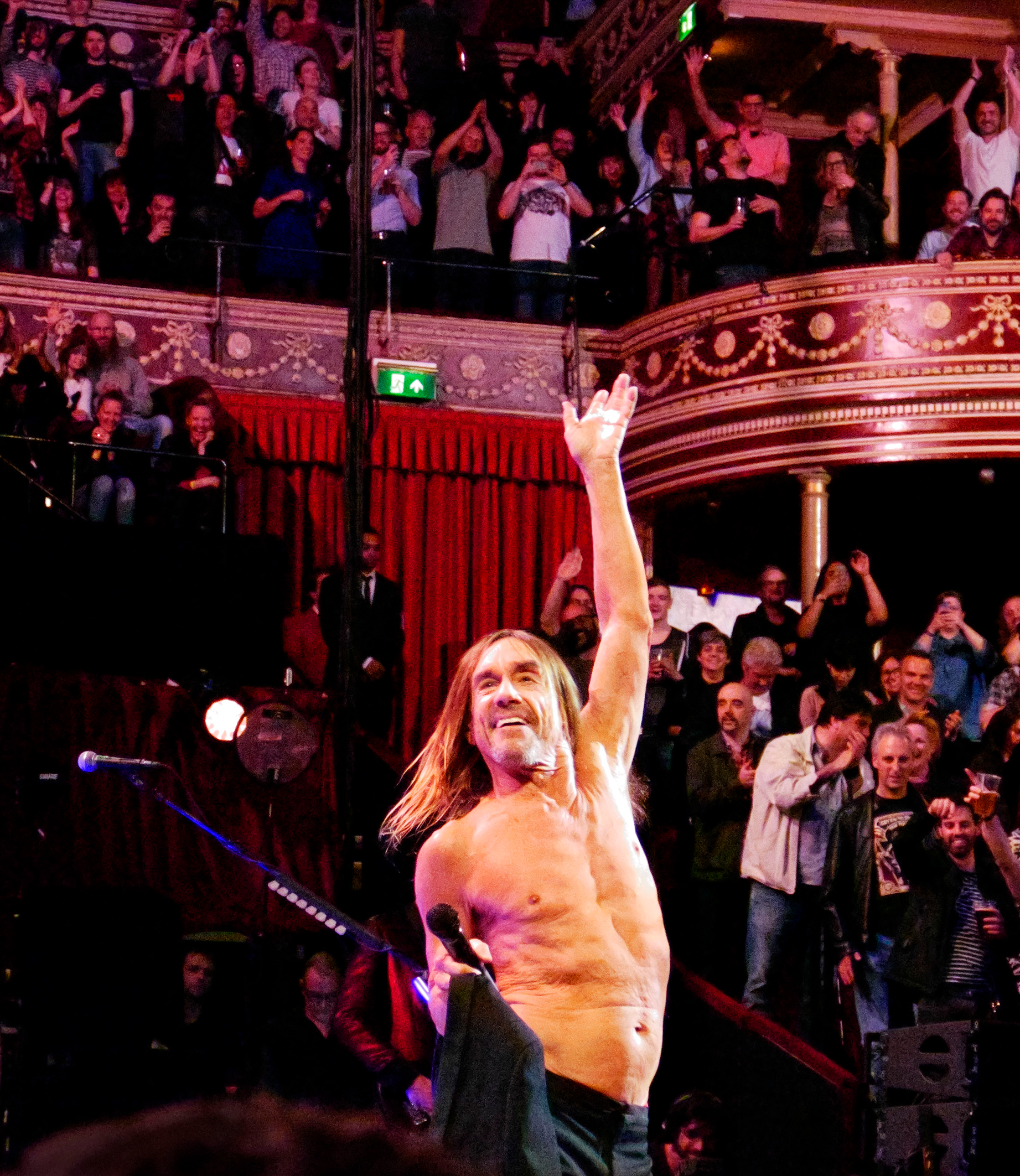 Shirtless male singer waves to a cheering crowd in an ornate concert hall with balconies and red curtains