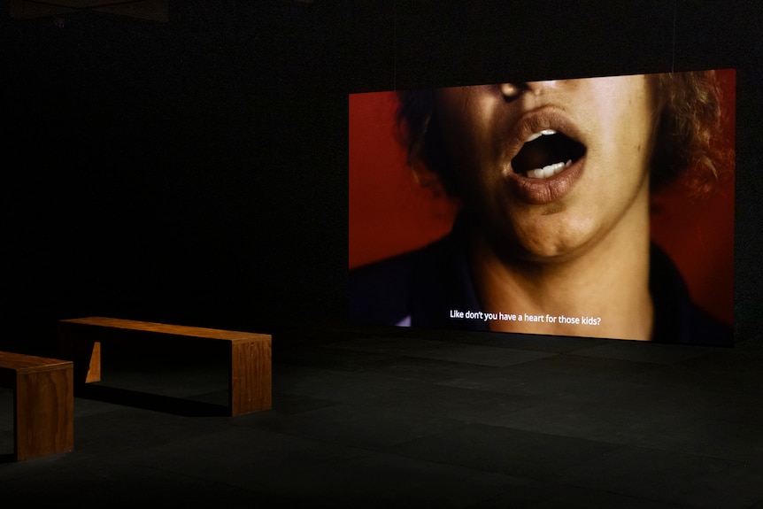 On a screen in a darkened gallery space is the image of the bottom half of a young woman of colour's face, mid-speech.