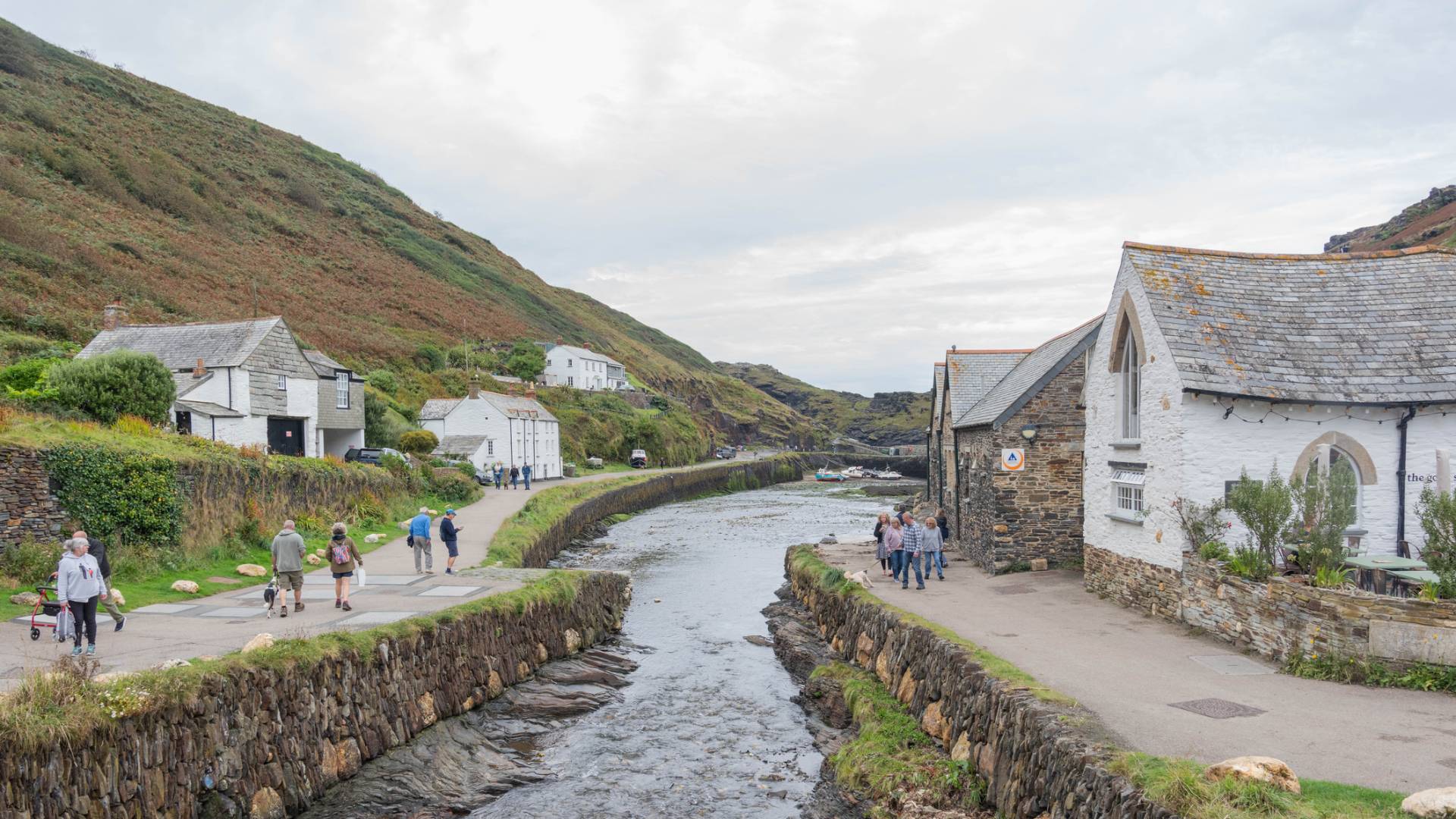 A narrow stream runs through a village with stone and white buildings on either side, set against a green hillside. People walk along the path beside the water under a cloudy sky.