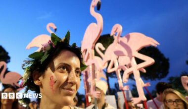 A man carries a large home-made puppet of a pink flamingo, flying above his head in a crowd of people protesting.