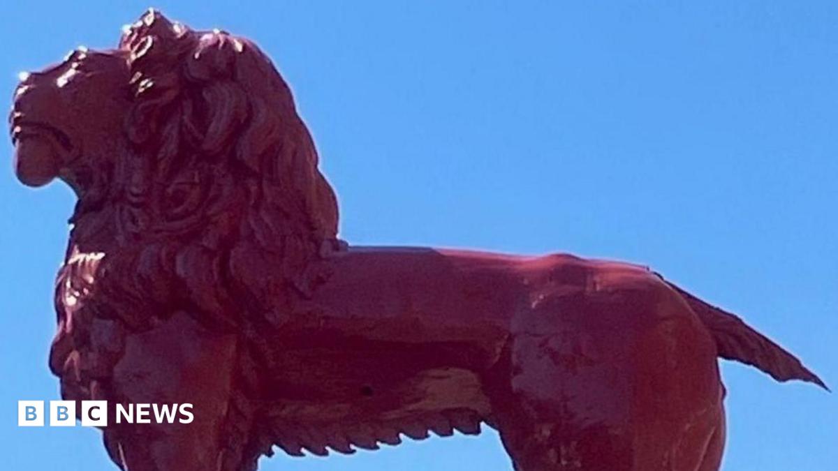 A wooden red lion sculpture stands atop a building, its mane and muscular form gleaming in the sunlight against a clear blue sky. Its tail has broken off.