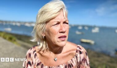 Melanie Leahy stands in front of Maldon harbour in Essex looking at the distance. She is wearing a pink and burgundy top with a silver heart locket around her neck. It is a sunny day. She has blonde, shoulder length hair. There are boats moored in the sea.