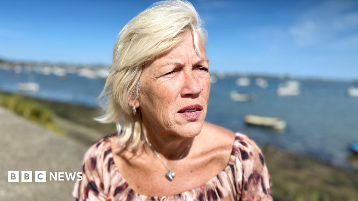 Melanie Leahy stands in front of Maldon harbour in Essex looking at the distance. She is wearing a pink and burgundy top with a silver heart locket around her neck. It is a sunny day. She has blonde, shoulder length hair. There are boats moored in the sea.