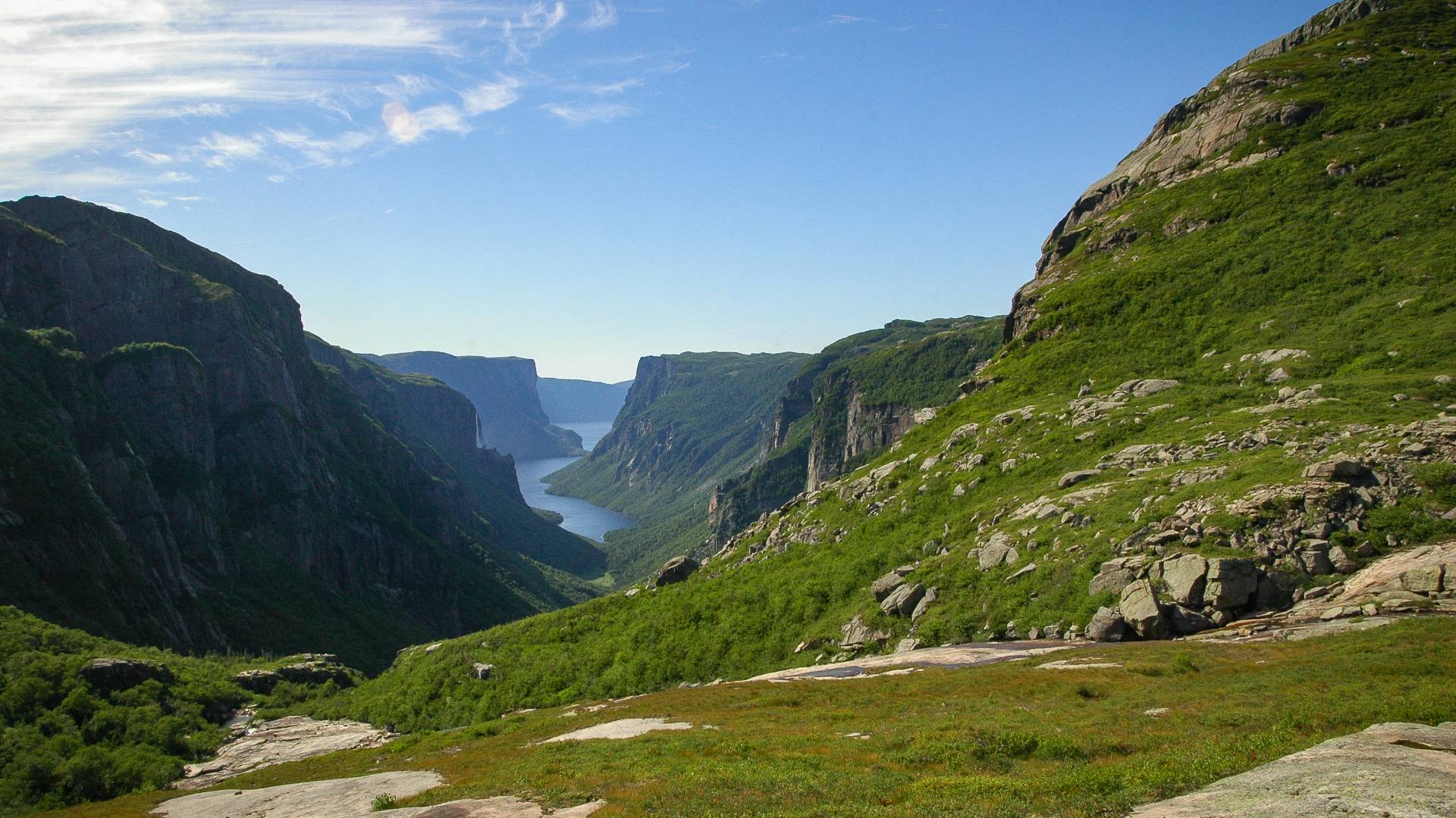 A scenic valley with steep, green hills and rocky slopes, opening to a narrow blue fjord stretching into the distance under a clear blue sky.