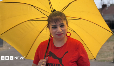 Amelia Lord is a white woman in her late 20s. She has shoulder length brown hair partly pulled back in a ponytail with frontpieces either side of her face. She has defined eyebrows and is wearing makeup, has a central nose ring and earrings, and is smiling at the camera. She wears a sleeveless black top. She is holding a pair of books and stands in front of a bookshelf with collections of books on it, including titles by Rebecca Yarros and the Harry Potter series by JK Rowling.