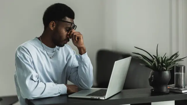 A tired-looking man rubs his eyes as he sits in front of his laptop, which is on a desk