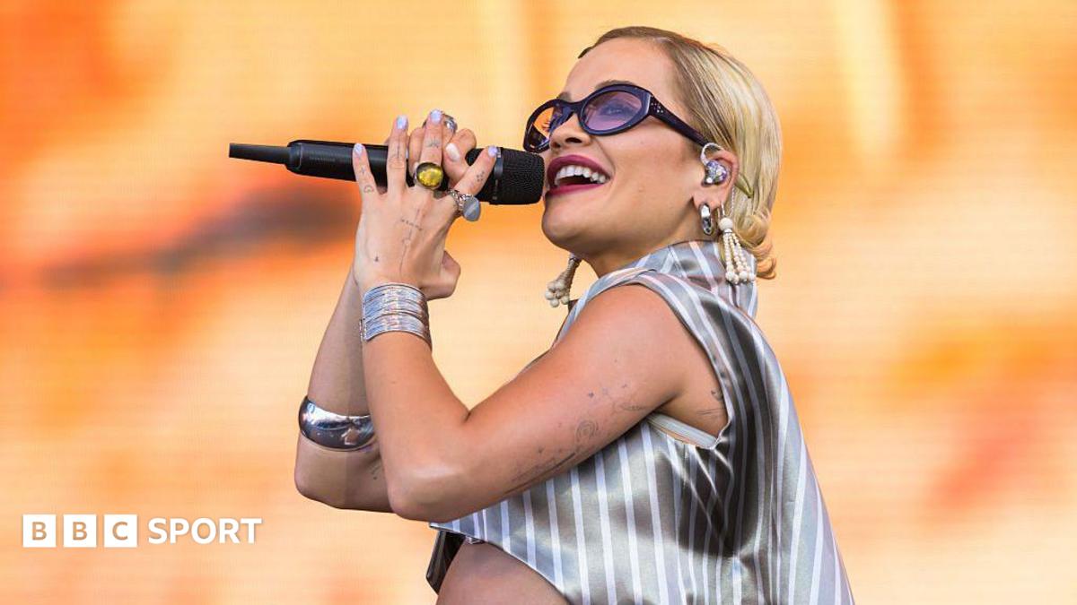 Rita Ora sings, holding a microphone and wearing a silver shirt and purple-tinted glasses