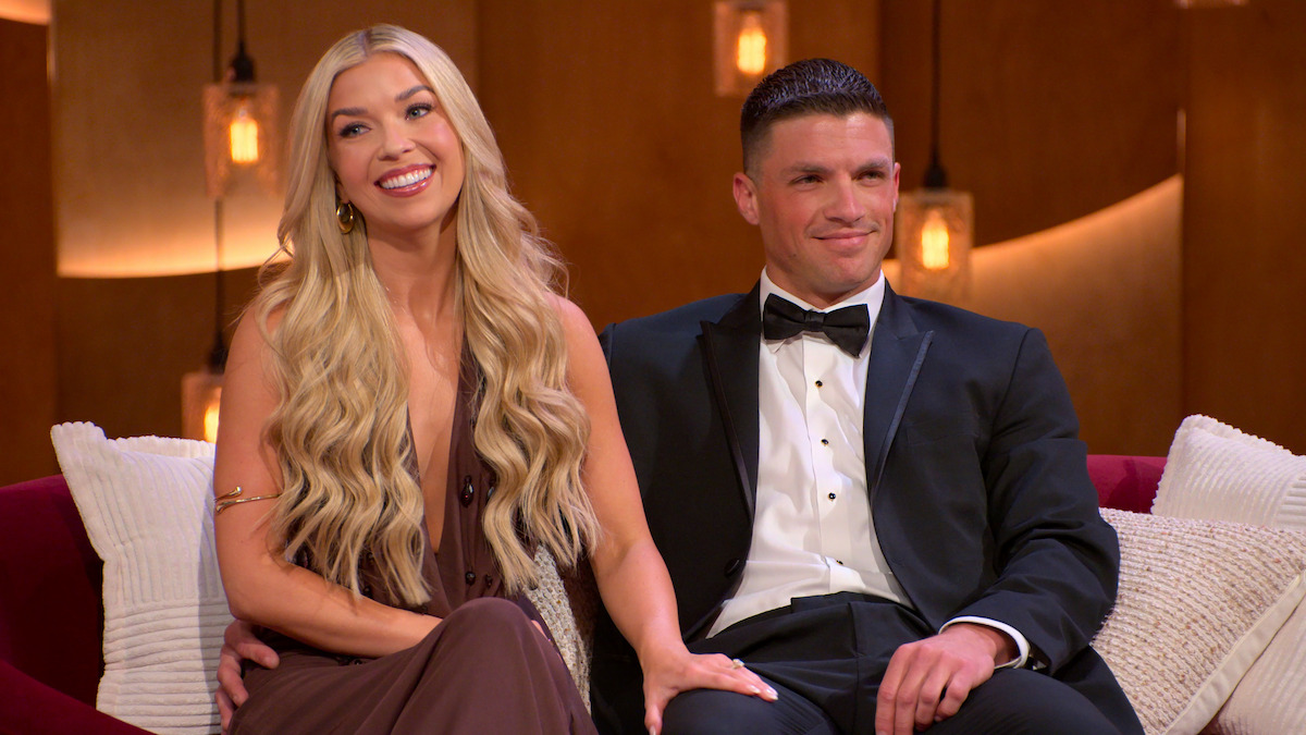 A smiling, blonde woman in a dress and a man in a tuxedo sit close together on a red couch in a warmly lit studio with hanging lights and pillows, appearing to participate in a formal interview or talk show.