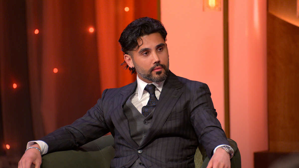 A man in a dark pinstripe suit sits on a green chair in a warmly lit room with brown and orange tones, looking attentively to the side. The environment suggests a formal or interview setting.