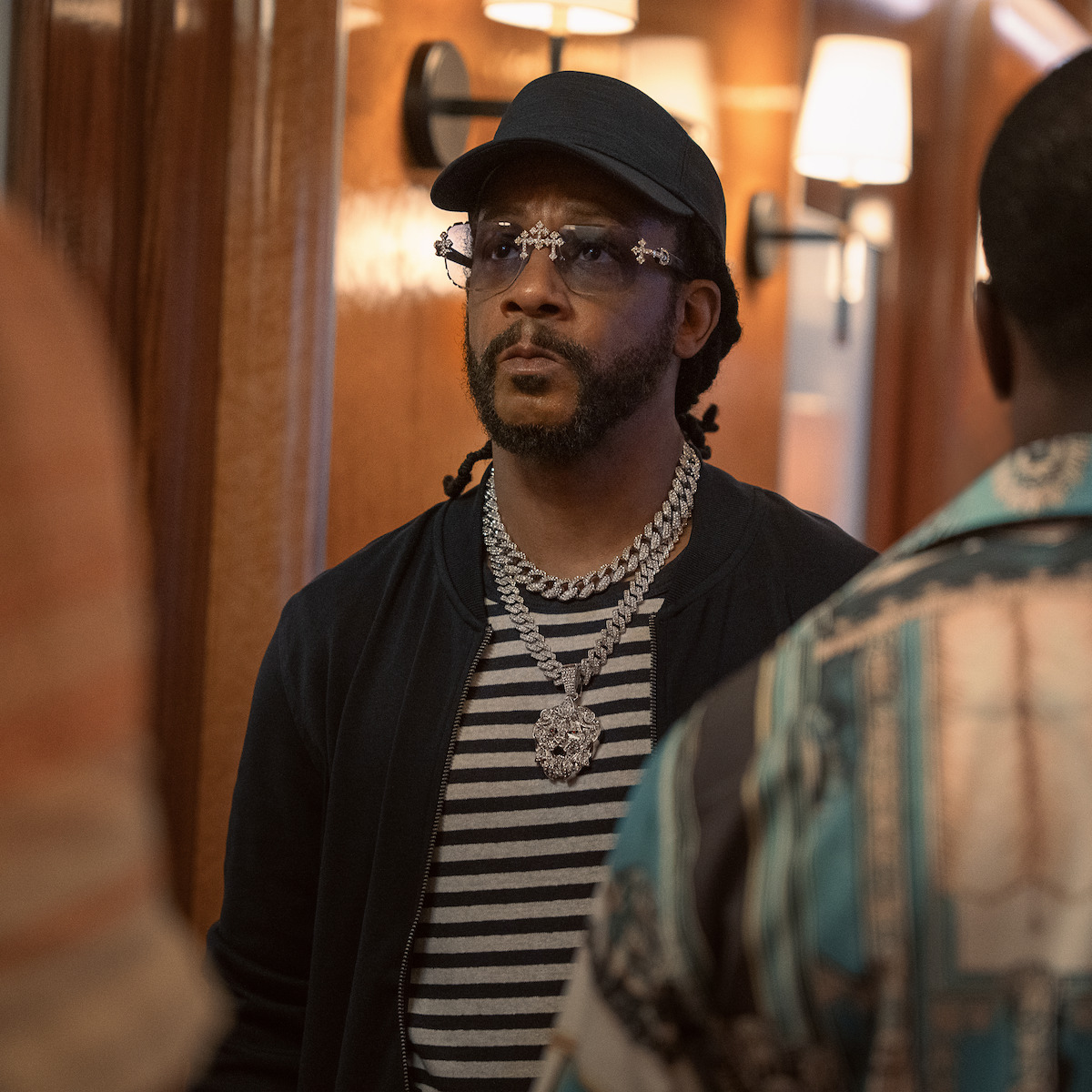 Man wearing sunglasses, cap, striped shirt, and thick chain necklace stands indoors in a warmly lit hallway, facing two other people whose backs are visible in the foreground.