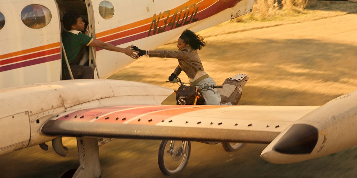 Person riding a motorcycle reaches out to grab another person's hand from an open plane door as the plane takes off on a dirt runway, creating a dramatic action scene outdoors.