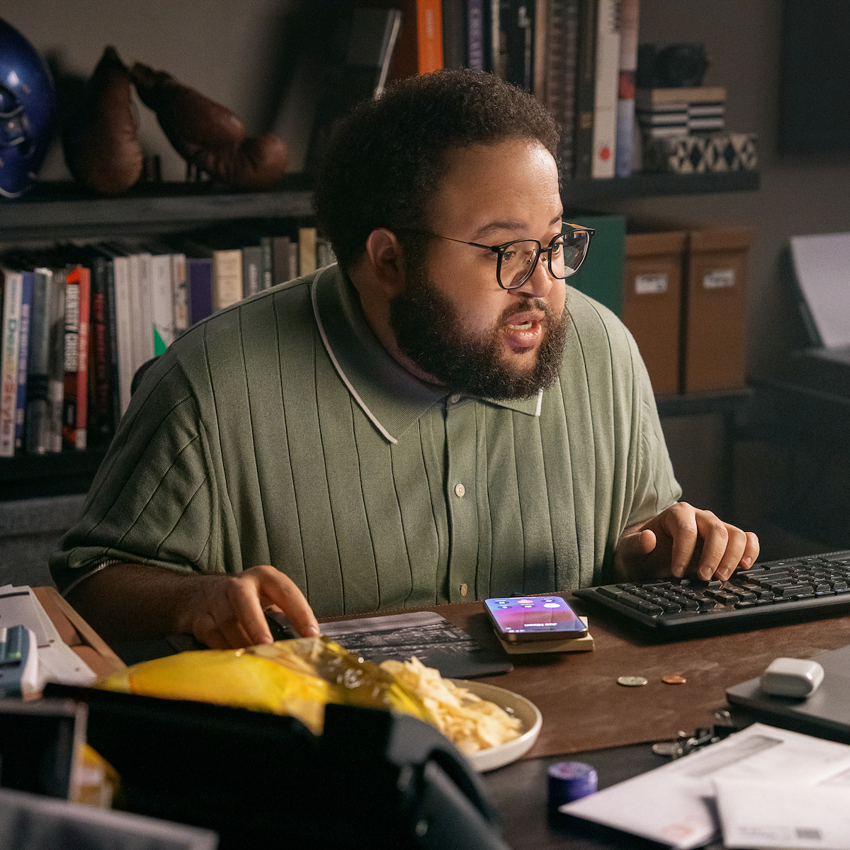 Zach Cherry as Barry in glasses sits at cluttered desk with food, smartphone, and computer keyboard, concentrating on monitor. Bookshelves and office supplies fill the background, suggesting a home or office workspace environment.