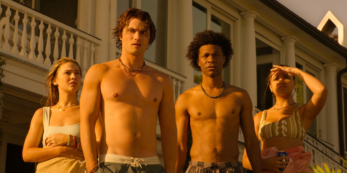 Four young adults stand outside a large house in warm, golden sunlight. Two are shirtless, and the group looks intently ahead, suggesting curiosity or concern. The setting feels summery and relaxed.