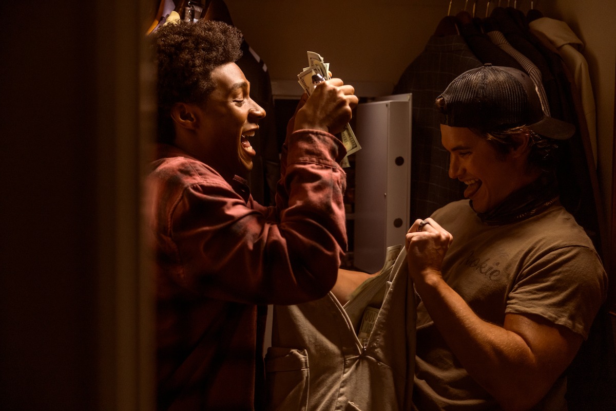 Two young men excitedly hold cash in a dimly lit closet, with clothes hanging in the background and an open safe visible behind them, suggesting they just found or stole the money.
