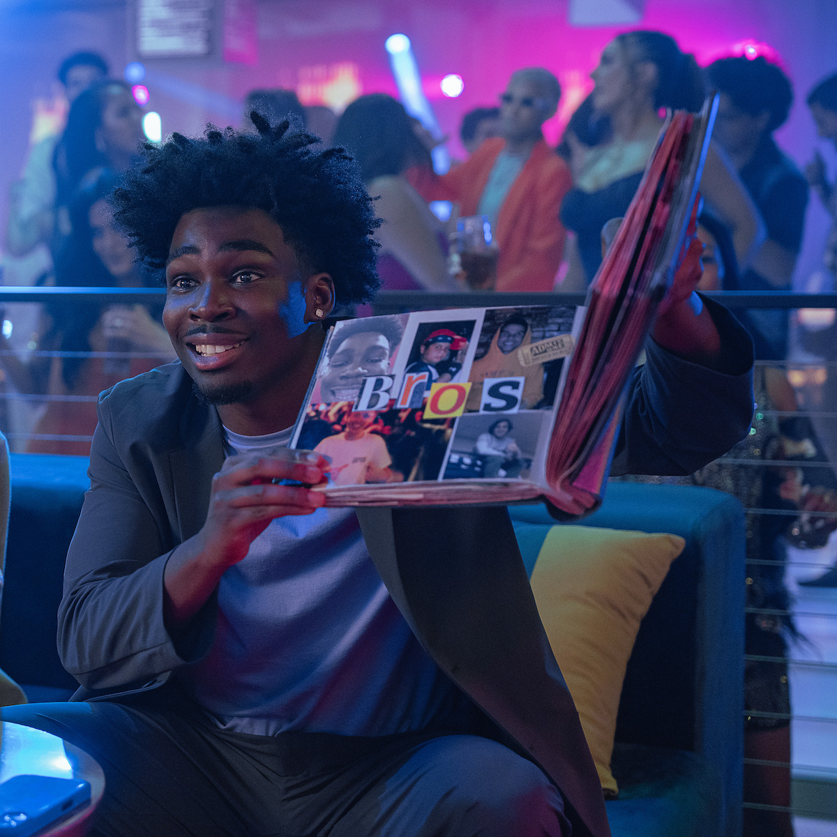Kam Patterson as Freshman sitting on a lounge chair in a lively, colorful nightclub holding up an open scrapbook with a “Bros” collage, with people dancing and socializing in the blurred background.