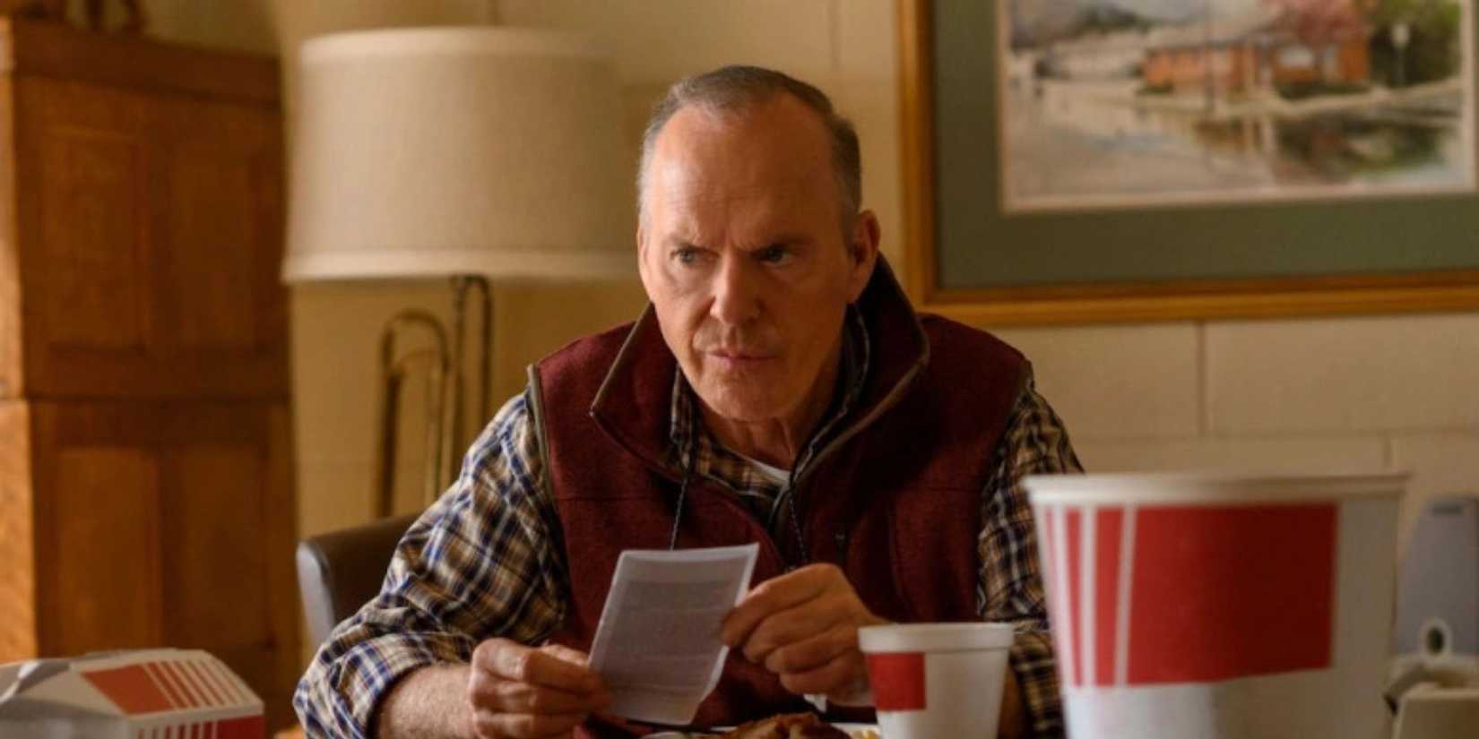 Michael Keaton sits at a table in a vest and checked shirt looking contemplative in Dopesick.