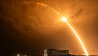 time lapse photo of an early morning rocket launch producing a bright streak arcing over a hangar and stretching beyond the clouds