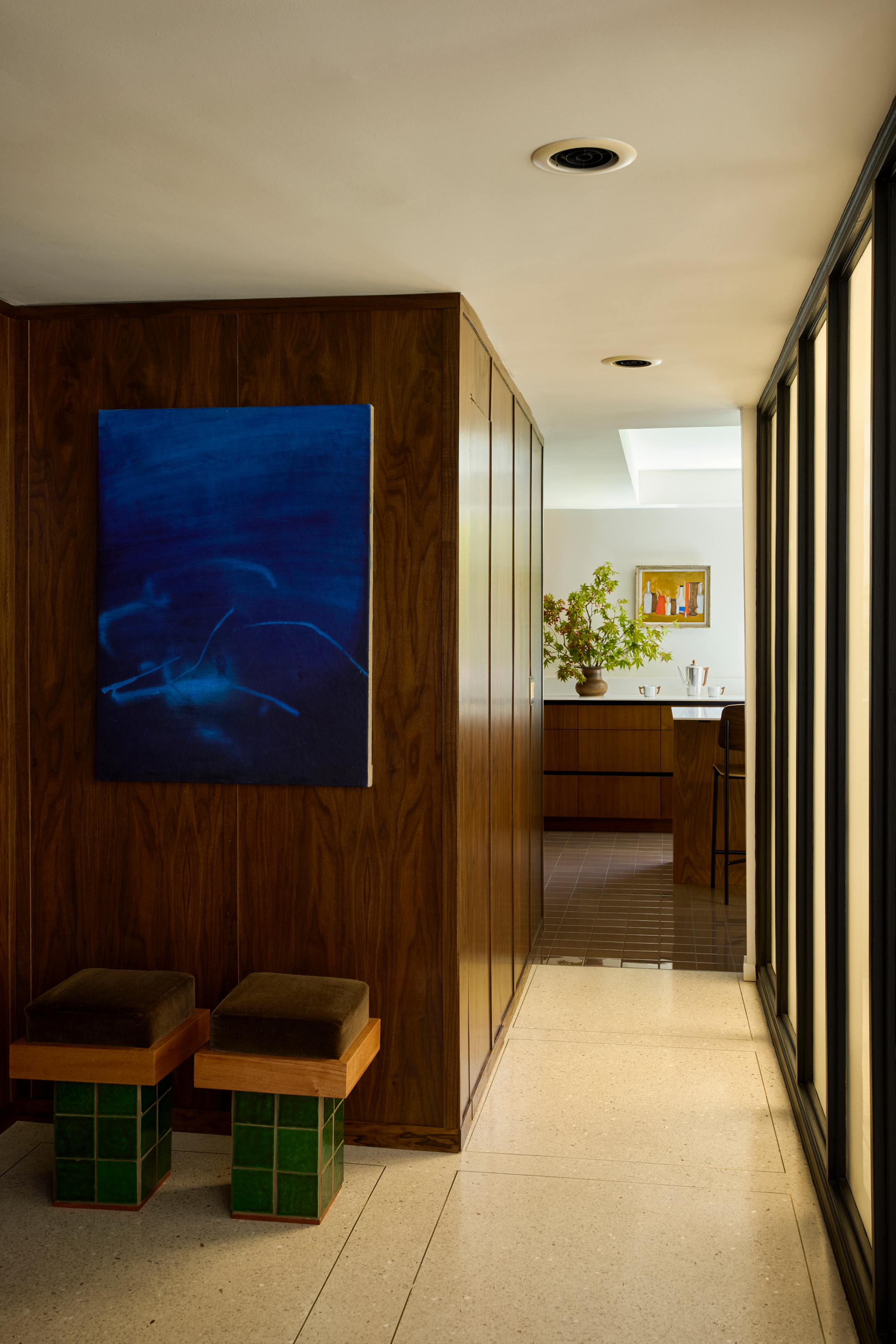 Small hallway between windows and wood joinery with stools in green tile and brown velvet upholstery and wood kitchen in the background