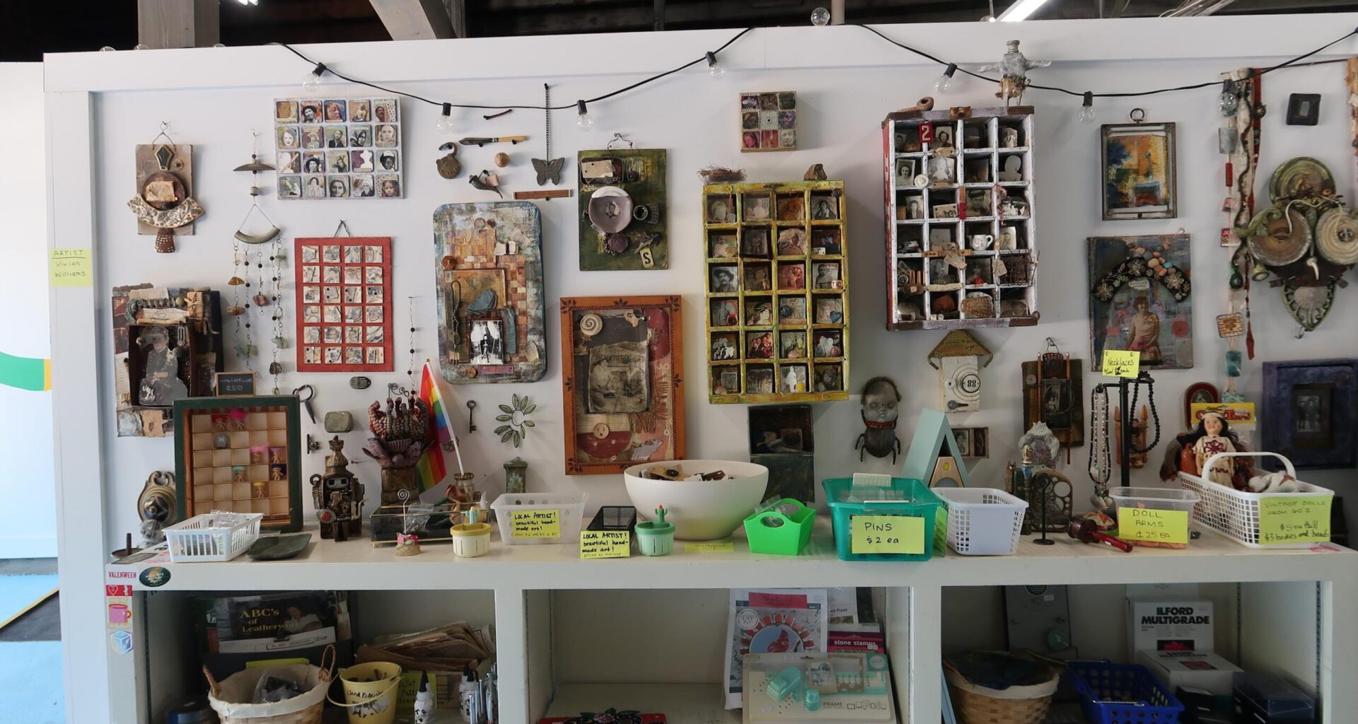 Shop display of handmade craft items: framed photos, jewelry, trinkets, and small boxes arranged on shelves with price tags and baskets beneath.