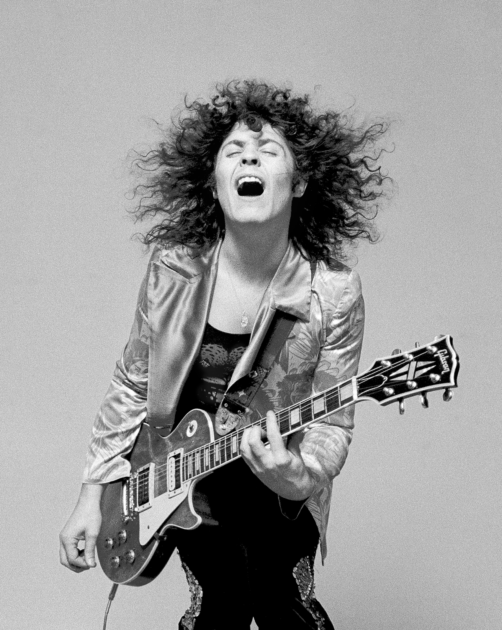 Black-and-white photo of a man with curly hair passionately playing an electric guitar, eyes closed, and mouth open