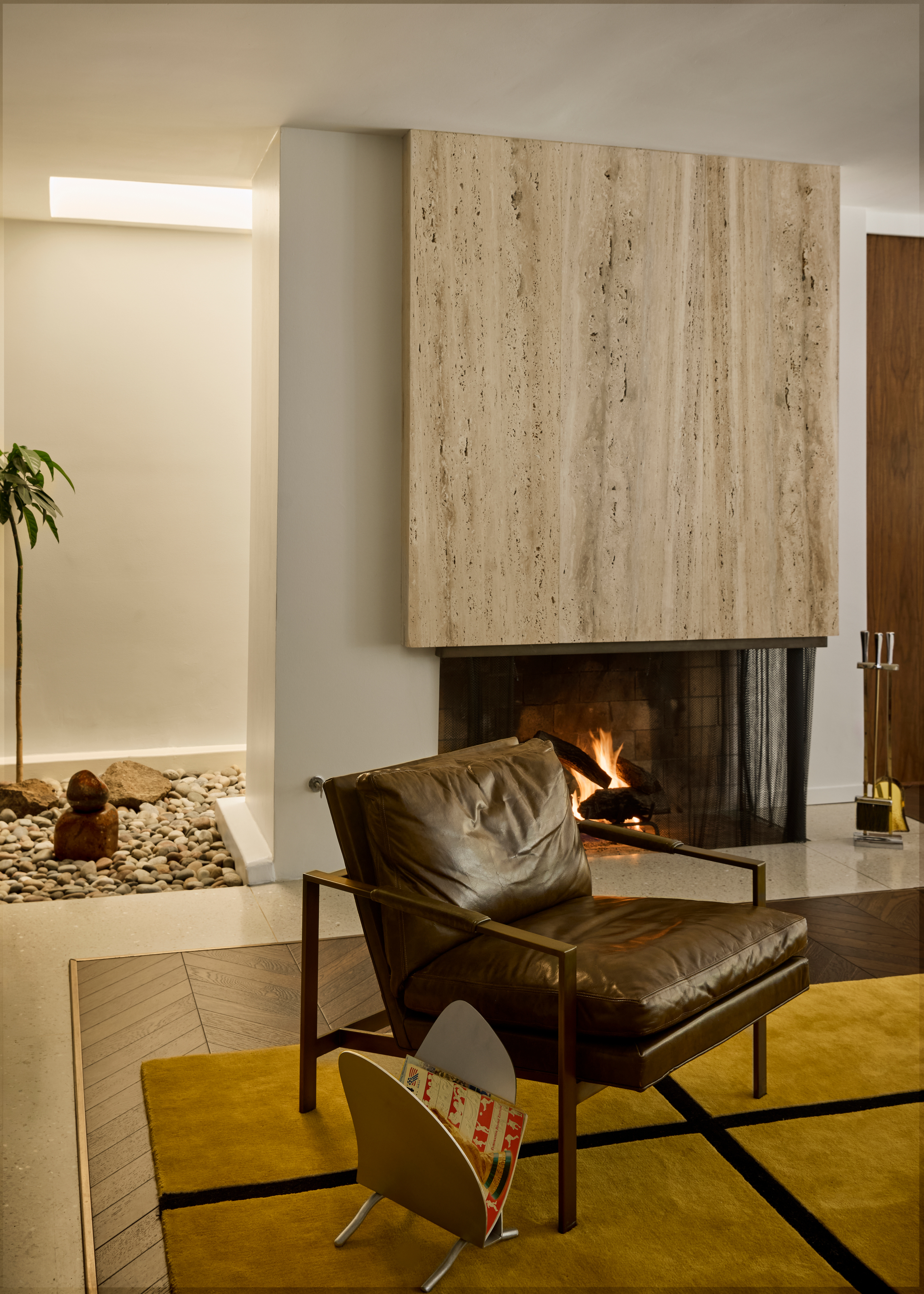 Travertine fireplace set into the wall behind a dark brown leather armchair, steel magazine rack and mustard and black-gridded rug