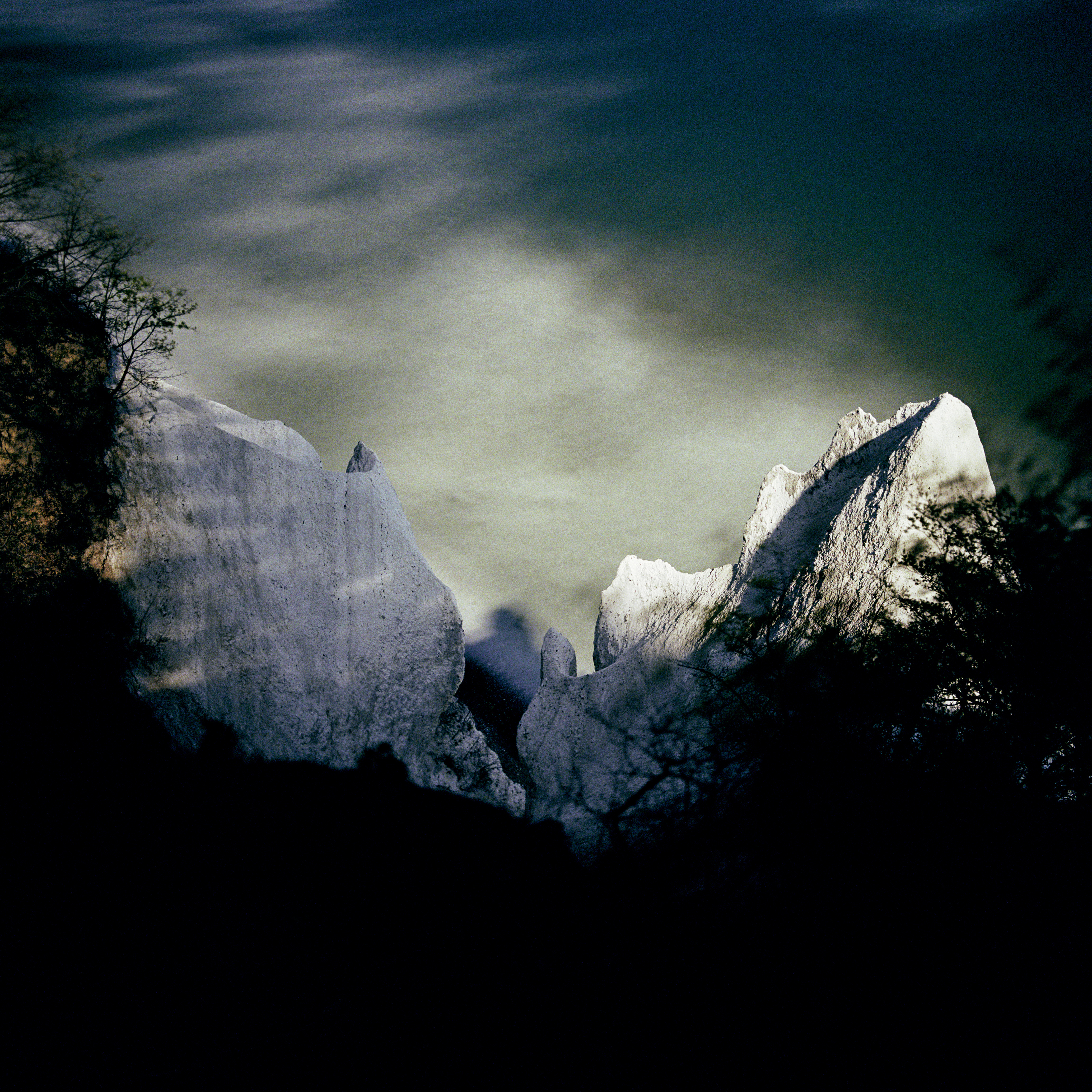A long-exposure photograph taken under full moonlight showing jagged, illuminated white cliffs surrounded by dark, silhouetted foliage and a hazy sky.