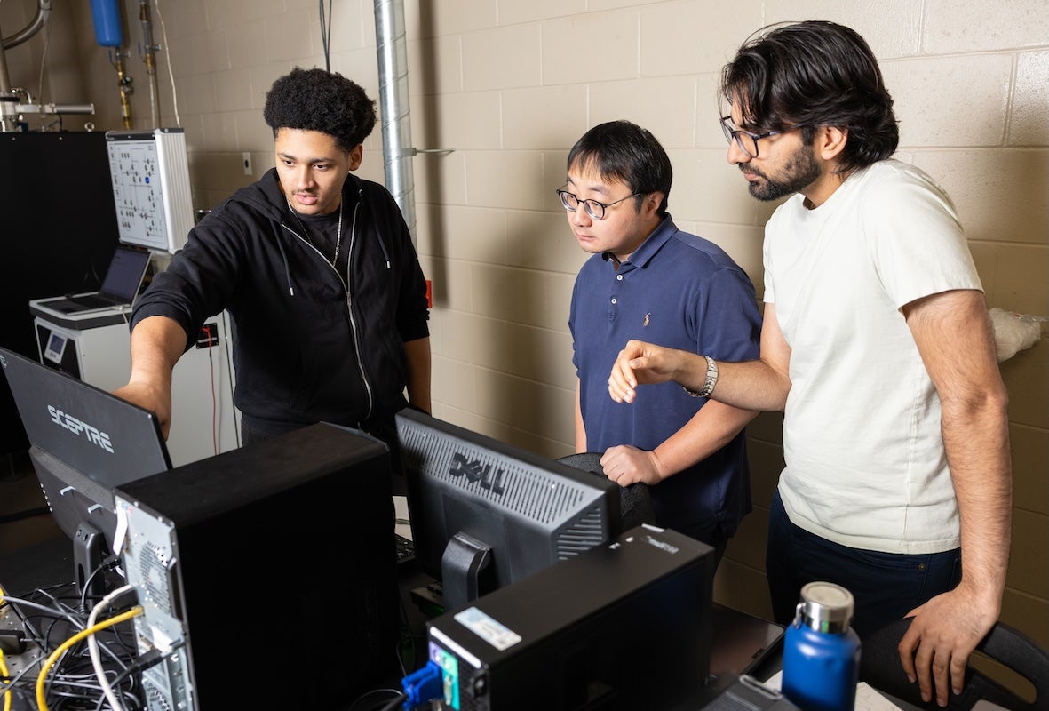 Three researchers gathered around computer monitors in a lab, one pointing at a screen while others watch, illustrating collaborative data review during experiments on superconducting and mechanical quantum systems.