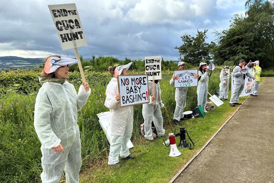 A line of people wearing white overalls and gannet masks stand in a line on a grassy area, holding up placards.