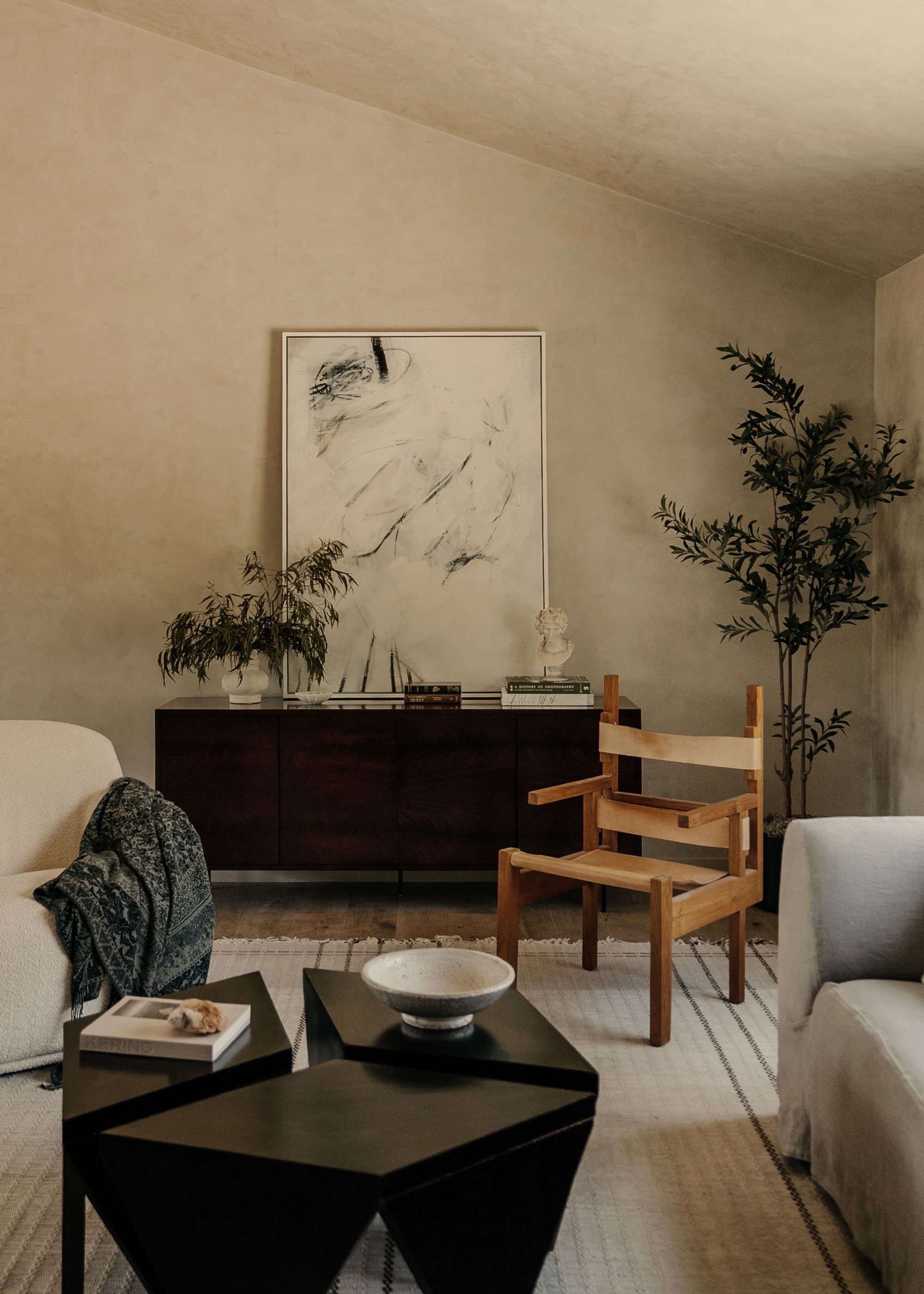 A minimalist living room with a potted tree, a vase of stems, a leaning painting, a wood accent chair, a gray sofa, a cream armchair, and a wood coffee table