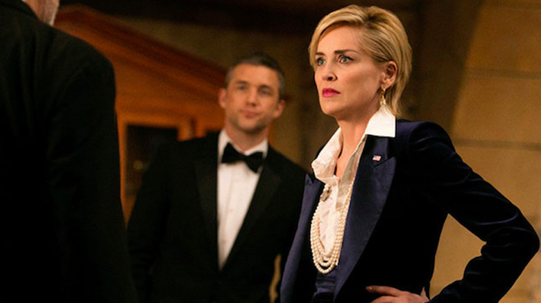 Sharon Stone as Natalie Maccabee with a serious expression in a navy blazer and pearls, looking at a man in the foreground, while Jeff Hephner as John Case stands behind her in a scene from 