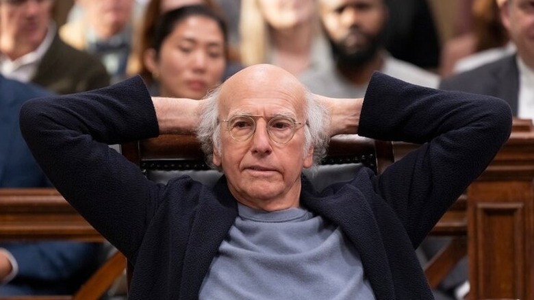 Larry David sitting at a defense table in a courtroom, leaning back with his hands clasped behind his head in a scene from 