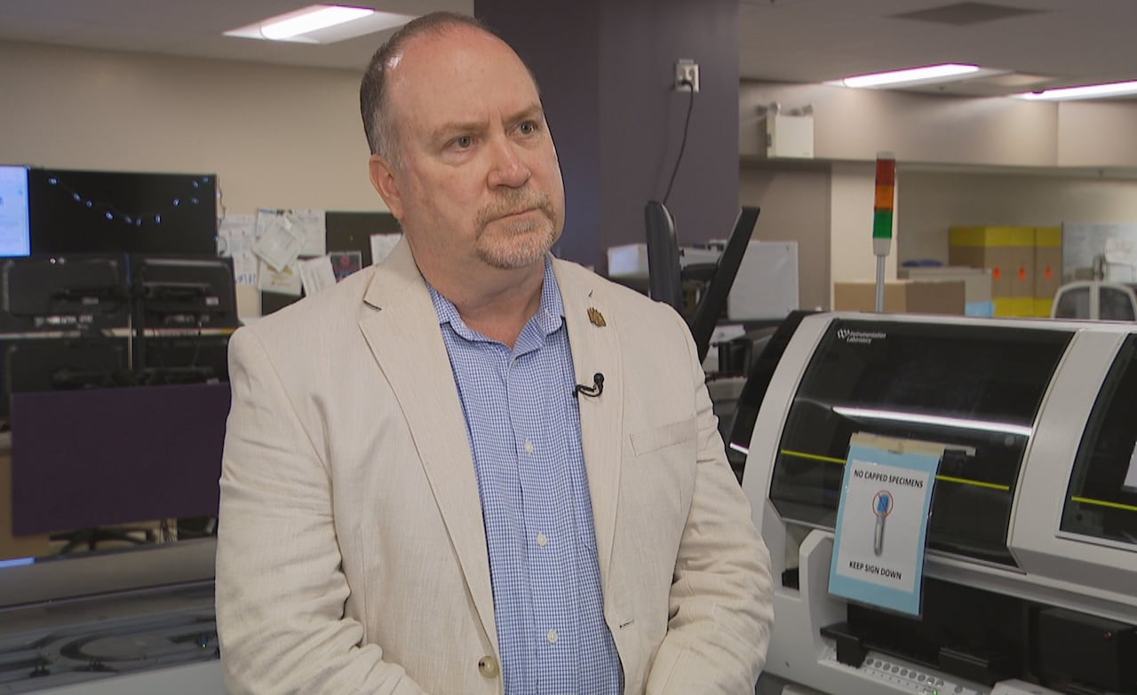 A man in a suit jacket stands in front of large lab equipment.