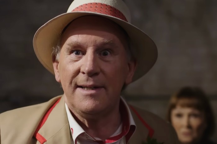 doctor-who-daleks Peter Davison as the Fifth Doctor in Doctor Who, wearing a cream straw hat and beige coat with red trim stands in a dim sci-fi interior.