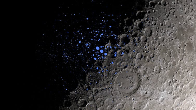 A close-up shot of the Moon's surface, with the left half covered in shadow, and the right half visible, with gray craters. Tiny blue dots in the center indicate permanently shadowed regions.