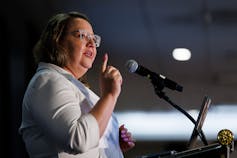 A woman wearing glasses and a white shirt gestures while speaking into a microphone