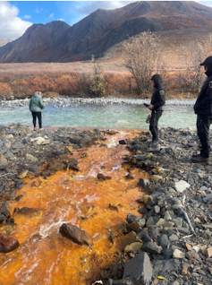 three people stand near a stream that has turned orange, it is flowing into another body of water. Mountains are in the background.