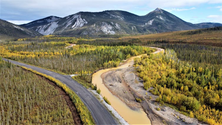 a beige colored river near a road in a forest area with a rocky mountain range in the distance