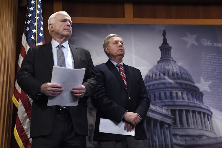 Two men in suits and ties stand on a stage with an American flag.